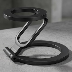 May include: A matte black, foldable phone stand. The stand features a circular base and a top ring to hold a phone. The stand is made of metal and has a modern, minimalist design. The text "FOR MASSAGE LABS PAT APPLIED FOR" is visible.