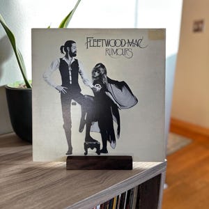 May include: A Fleetwood Mac "Rumours" vinyl record album cover featuring a black and white image of two people. The album is displayed on a dark wooden stand. The album cover has the band's name and album title.