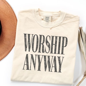 May include: Cream-colored t-shirt with the words "WORSHIP ANYWAY" printed in dark gray, distressed lettering. The shirt has a crew neck and a small tag that says "Comfort Colors". A gold necklace with a small cross pendant is visible.