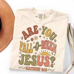 May include: A cream-colored t-shirt featuring a graphic print with the text "ARE YOU FALL-O-WEEN JESUS?" and "MATTHEW 4:19" in wavy, multi-colored lettering. The design includes small flower, star, and sun icons in shades of orange, yellow, and green.