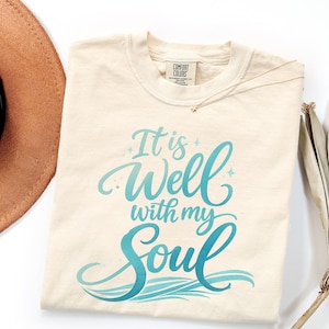 May include: Cream-colored t-shirt with the text "It is Well with my Soul" in a teal script. The shirt has a crew neck and a Comfort Colors tag. A gold necklace and a brown hat are partially visible.