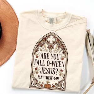May include: A cream-colored t-shirt featuring a graphic of a gothic-style arched window frame. Inside the frame, the text reads "ARE YOU FALL-O-WEEN JESUS? MATTHEW 4:19" above a small pumpkin with a cross on top, surrounded by scattered autumn leaves.