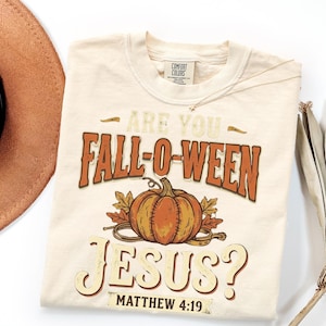May include: A cream-colored t-shirt featuring a graphic with an orange pumpkin, autumn leaves, and the text "ARE YOU FALL-O-WEEN JESUS?" in a distressed font, along with the scripture reference "MATTHEW 4:19" printed at the bottom of the design.