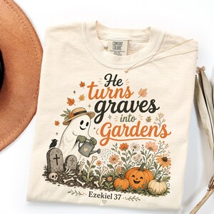May include: A cream-colored t-shirt featuring a graphic of a ghost wearing a straw hat and holding a watering can. The text reads "He turns graves into Gardens" and "Ezekiel 37". The design includes tombstones, bones, pumpkins, and flowers.