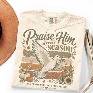 May include: A cream-colored t-shirt featuring a graphic of a flying dove holding an olive branch. The text reads Praise Him in every season and His praise outlives every season. The design includes pumpkins, sunflowers, and autumn leaves.
