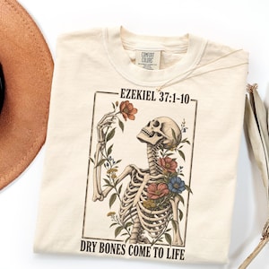 May include: A cream-colored t-shirt featuring a graphic of a skeleton surrounded by colorful flowers. The text "EZEKIEL 37:1-10" appears above the illustration, and the text "DRY BONES COME TO LIFE" is printed below the rectangular frame.