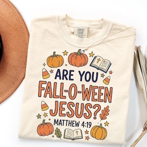 May include: A cream-colored t-shirt featuring a graphic print with the text "ARE YOU FALL-O-WEEN JESUS? MATTHEW 4:19". The design includes orange pumpkins, candy corn, open bibles with a cross, small stars, and autumn leaves in a retro style.