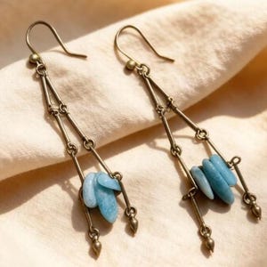 May include: A pair of dangle earrings with a vintage aesthetic. The earrings feature linked bronze-coloured metal pieces, with a cluster of light blue stones at the bottom. The earrings are suspended from a bronze-coloured hook.