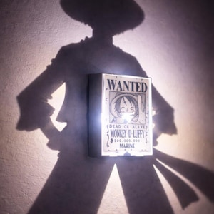 以下が含まれることがあります： モンキー・D・ルフィの画像が描かれた「WANTED」ポスターが光る長方形の看板。質感のある壁に映る看板の影は、帽子とコートを着た人物を描いています。