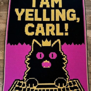 Pode incluir: Uma manta de crochê retangular com fundo preto e o texto amarelo I AM YELLING, CARL! sobre um gráfico de um gato preto com coroa amarela, olhos e boca rosa choque, sentado em frente a um teclado de computador amarelo e preto.