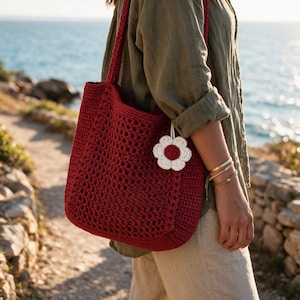 Peut inclure: Un sac fourre-tout en crochet rouge foncé présentant un motif à mailles ouvertes texturé sur le panneau avant et une bandoulière en crochet solide. Une petite breloque en forme de fleur crochetée blanche et rouge est suspendue sur le côté.