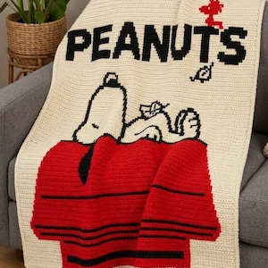 May include: A cream-colored crochet blanket featuring the black text PEANUTS at the top. Below the text, a white Snoopy character is depicted lying on a red doghouse with black horizontal stripes. A small red Woodstock figure is also visible.