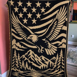 Puede incluir: Una manta de ganchillo rectangular con un esquema de color negro y beige. El diseño muestra un águila grande en vuelo sobre una cordillera y un paisaje forestal, con un patrón de estrellas dispuestas en la parte superior del tejido.