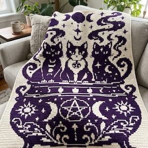Könnte beinhalten: Eine rechteckige Häkeldecke in Dunkelviolett und Creme. Das Design zeigt drei Katzen, die hinter einem großen Kessel sitzen, der mit einem zentralen Pentagramm, Mondsicheln, Sonnensymbolen und wirbelnden Rauchmustern verziert ist.