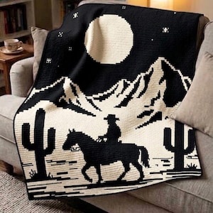 May include: A black and cream crochet blanket featuring a desert landscape design. The pattern displays a silhouette of a person riding a horse, two saguaro cacti, a large full moon, and small stars against a dark night sky background.