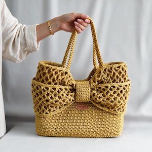 Pode incluir: Uma bolsa de crochê amarelo-mostarda com um grande laço na parte frontal. A bolsa possui uma base densa e texturizada, além de um padrão de rede vazado na parte superior e no laço. Uma pequena etiqueta retangular Handmade está fixada.
