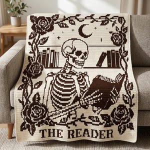 May include: A cream-colored crochet blanket featuring a dark brown graphic design of a skeleton reading a book in a library setting. The border is decorated with roses and vines, with a crescent moon above and the text THE READER at the bottom.