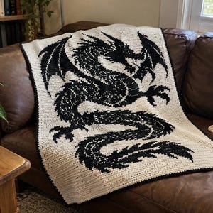 May include: A rectangular crochet blanket featuring a large, detailed black dragon silhouette centered against a cream-colored background. The design is created with a textured stitch pattern, and the entire blanket is finished with a thin black border.