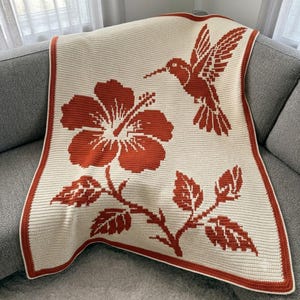 Puede incluir: Una manta de ganchillo color crema con una gran flor de hibisco color naranja óxido, un colibrí en vuelo y tallos con hojas. La manta está rematada con un borde a juego en color naranja óxido que enmarca toda la pieza textil rectangular.