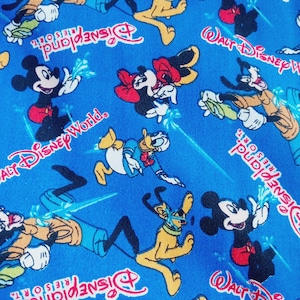 Puede incluir: Tela azul brillante con un patrón repetido de personajes de Disney, incluyendo Mickey Mouse, Minnie Mouse, Donald Duck y Pluto. Las palabras "Walt Disney World" y "Disneyland Resort" también son visibles en rojo y blanco. Los personajes están en poses de acción.