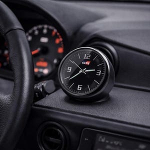Pode incluir: Um relógio analógico de carro preto e redondo com um mostrador minimalista apresentando números brancos nas posições de 3, 6, 9 e 12 horas. O centro do mostrador exibe as letras GR em um logotipo vermelho e branco. Ponteiros brancos finos.