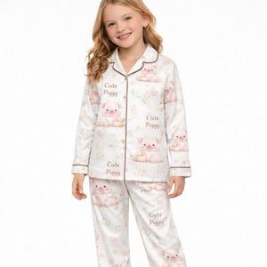 May include: White pajama set with a repeating pattern of pink pigs and floral accents. The set includes a button-down top and matching pants. The text "Cute Piggy" is printed on the top and pants.
