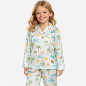 Op de afbeelding: Witte pyjamaset met een tropische print met palmbomen, zonnen, schelpen en zonnebrillen. De top met lange mouwen heeft een knoopsluiting en een kraag. De bijpassende broek maakt de set compleet.