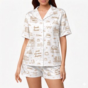 May include: White pajama set with a repeating pattern of books, coffee cups, and glasses in a light brown color. The shirt has the text "book lover" and "just one more chapter". The shorts have the same text. The set is made of a soft, silky material.