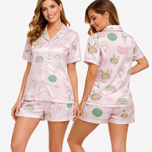 May include: Pink satin pajama set with a whimsical pattern of rainbows, flowers, and other playful shapes. The set includes a short-sleeved button-up shirt and matching shorts, both trimmed with a light green piping.