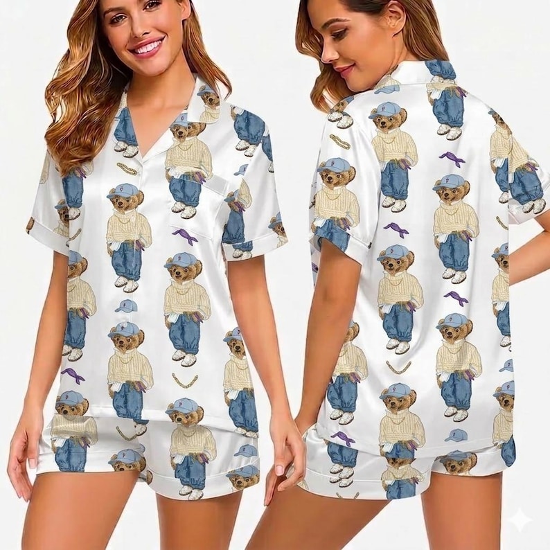 Puede incluir: Conjunto de pijama de sat&eacute;n blanco con un estampado repetido de un oso de peluche con gorra azul, su&eacute;ter y vaqueros. El conjunto incluye una camisa de manga corta con botones y pantalones cortos a juego. El pijama tiene un corte holgado.