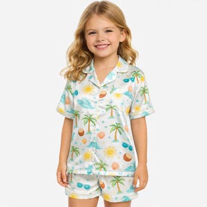 Op de afbeelding: Witte kinderpyjamaset met een tropische print van palmbomen, zonnen, golven en zonnebrillen. De top met korte mouwen en de shorts zijn gemaakt van een zijdeachtig materiaal.