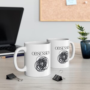 May include: Two white ceramic mugs with the word "OBSESSION" printed in black, along with a black scribble design. The mugs have a curved handle and are sitting on a light-colored surface.
