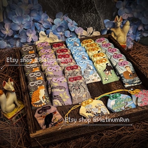 Puede incluir: Una bandeja de madera llena de coloridos amuletos omamori japoneses. Los amuletos presentan varios diseños, incluyendo gatos, serpientes e ilustraciones. El fondo incluye flores azules y figuras decorativas de zorro.