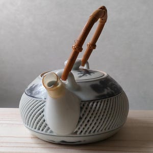 May include: A white and blue ceramic teapot with a bamboo handle. The teapot has a round body with a lattice design and a spout. The handle is made of two curved bamboo pieces. The lid has a floral design.