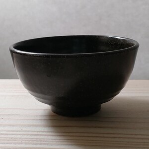 以下が含まれることがあります： わずかにテクスチャ加工された表面を持つ、濃い茶色のセラミックボウル。ボウルは丸みを帯びた形状で、足付きのベースが付いています。内側は濃い色で、明るい色の木製の表面に置かれています。
