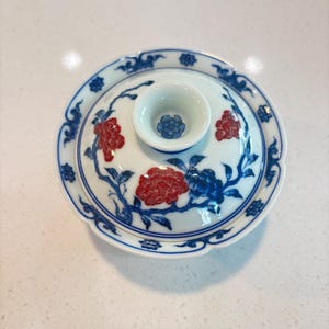 May include: A white ceramic lidded bowl with a blue and red floral design. The bowl features a small central cup and is decorated with blue vines and red flowers. The rim of the bowl is also decorated with blue patterns.