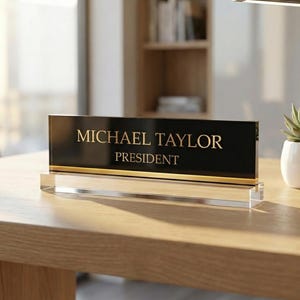 May include: A black desk nameplate with gold lettering that reads "MICHAEL TAYLOR PRESIDENT". The nameplate is on a clear acrylic base and has a gold stripe along the bottom edge. The nameplate is on a wooden desk.
