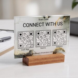 May include: A rectangular acrylic sign with a white and gold marble pattern, featuring the text CONNECT WITH US and three QR codes labeled Cash App, Venmo, and Instgram. The sign is held upright by a rectangular wooden base stand.
