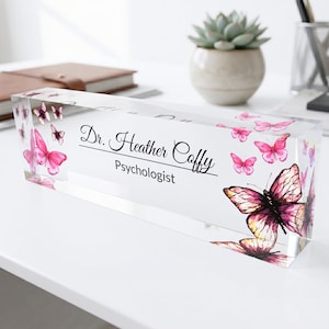Puede incluir: Una placa de nombre para escritorio rectangular de acrílico transparente con ilustraciones de mariposas rosas y texto impreso en negro que dice Dr. Heather Coffy Psychologist. El diseño incluye varias mariposas de distintos tamaños.