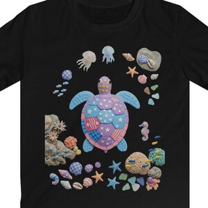 May include: Black t-shirt with a vibrant sea turtle illustration. The turtle's shell is a patchwork of blue, pink, and purple. The design includes jellyfish, starfish, and seashells, creating a marine theme.