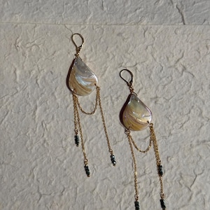 May include: A pair of drop earrings featuring teardrop-shaped iridescent white and gold marbled pendants. Each earring has gold-tone lever-back hooks, delicate gold chains draped in a loop, and small dark grey beads at the ends of the chains.