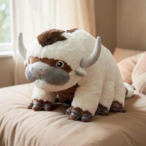 Pode incluir: Um brinquedo de pel&uacute;cia de uma criatura branca e fofa com manchas marrons, focinho cinza e chifres grandes e curvos. O brinquedo tem quatro patas com cascos listrados em marrom e azul, e est&aacute; em uma cama.