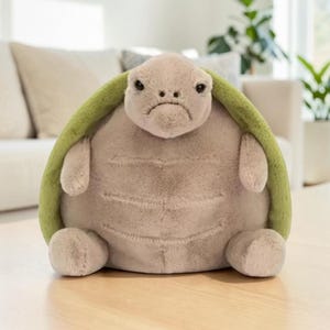 Puede incluir: Un peluche de tortuga con un caparazón verde claro y una expresión triste. El cuerpo de la tortuga es de color gris claro y suave, con extremidades pequeñas y rechonchas. El juguete está diseñado para ser tierno y probablemente está hecho de una tela suave.
