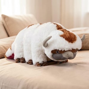 Pode incluir: Um brinquedo de pel&uacute;cia em forma de bis&atilde;o, com pelo branco e manchas marrons na cabe&ccedil;a e nas costas. O brinquedo tem pequenos chifres cinzentos, patas marrons e um rosto cinzento. Est&aacute; sobre uma superf&iacute;cie bege.