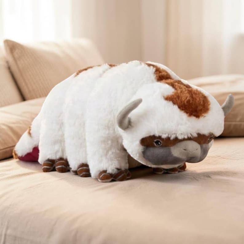 Pode incluir: Um brinquedo de pel&uacute;cia em forma de bis&atilde;o, com pelo branco e manchas marrons na cabe&ccedil;a e nas costas. O brinquedo tem pequenos chifres cinzentos, patas marrons e um rosto cinzento. Est&aacute; sobre uma superf&iacute;cie bege.