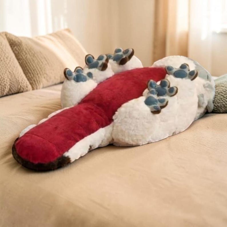 Pode incluir: Uma almofada em forma de p&eacute;, vermelha e branca, com almofadas azuis. A almofada tem uma sola vermelha e uma parte superior branca e fofa, projetada para se parecer com um p&eacute; gigante. Descansa sobre uma cama com roupa de cama bege.