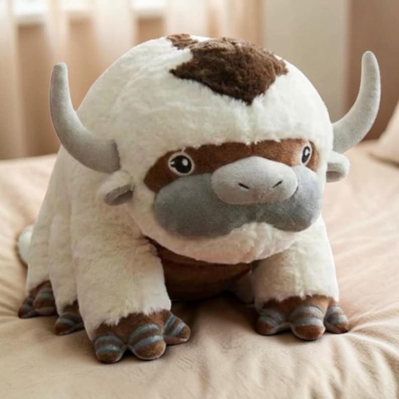 Pode incluir: Um brinquedo de pel&uacute;cia de uma criatura semelhante a um bisonte, principalmente branco com detalhes marrons e cinza. O brinquedo tem chifres grandes e curvos, um corpo arredondado e tra&ccedil;os faciais detalhados. Os p&eacute;s da criatura t&ecirc;m detalhes listrados.