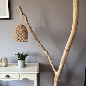 Könnte beinhalten: Eine einzigartige Stehlampe aus einem natürlichen Ast. Die Lampe hat einen gewebten Lampenschirm aus Rattan, der an einem kleineren Ast hängt. Der Sockel ist ein dunkelbrauner Metalleimer mit einem Jutesack.