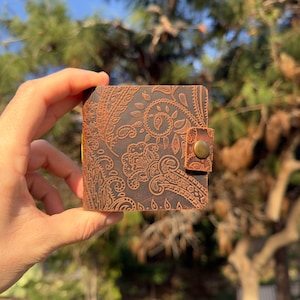 May include: A brown leather wallet with an embossed paisley design. The wallet has a snap closure with a brass button. The wallet is held in a hand against a blurred green background.