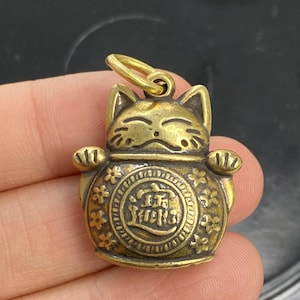May include: A small, gold-colored Maneki-neko charm. The charm features a smiling cat with raised paws and a round body. The cat has a circular design with Asian characters. The charm has a small loop at the top for hanging.